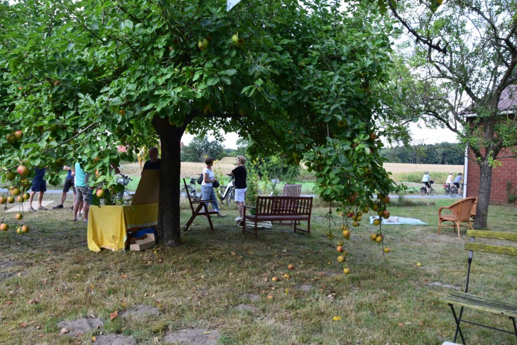 Ein großer Apfelbaum, der Apfel trägt, steht auf einer Wiese auf der Fallobst liegt. Unter dem Baum steht ein Tisch mit einer gelben Tischdecke, sowie Holzbänke und Stühle. Zwei Menschen unterhalten sich. Im Hintergrund liegt ein Kornfeld. Auf der Straße davor fahren Menschen mit dem Fahrrädern. Am rechten Bildrand ist die Ecke eines Hauses aus roten Backsteinen zu erkennen.