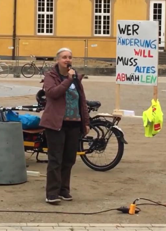 S4F-Mitglied Carola Meyer spricht beim Klimastreik vor dem Schloss durch ein Mikrofon zu den anwesenden Demonstrant:innen. Hinter ihr ist ein Plakat mit der Aufschrift „Wer Änderung will, muss Altes abwählen“ zu sehen.