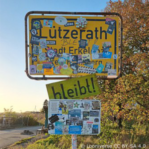 Ortsschild von Lützerath mit vielen Stickern drauf. darunger ein gelbes Schild mit schwarzer Aufschrift "bleibt!"