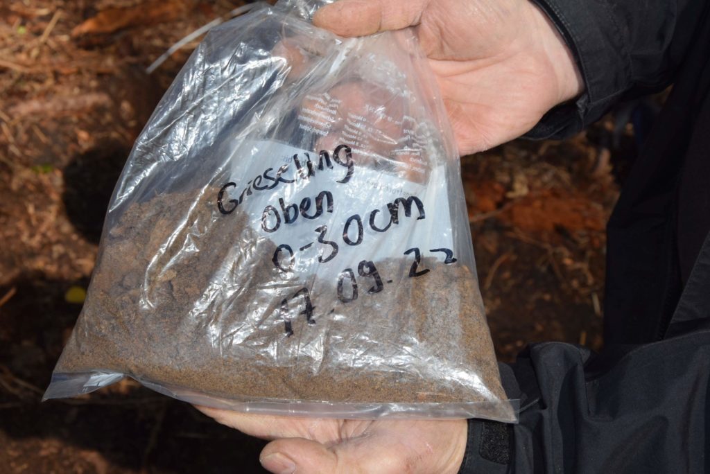 Zwei Hände halten eine beschriftete Plastiktüte. "Grieseling, Oben, 0-30cm, 17.09.22" In der Tüte befindet sich die Bodenprobe aus hellbraunem, sandigem Bodenmaterial.