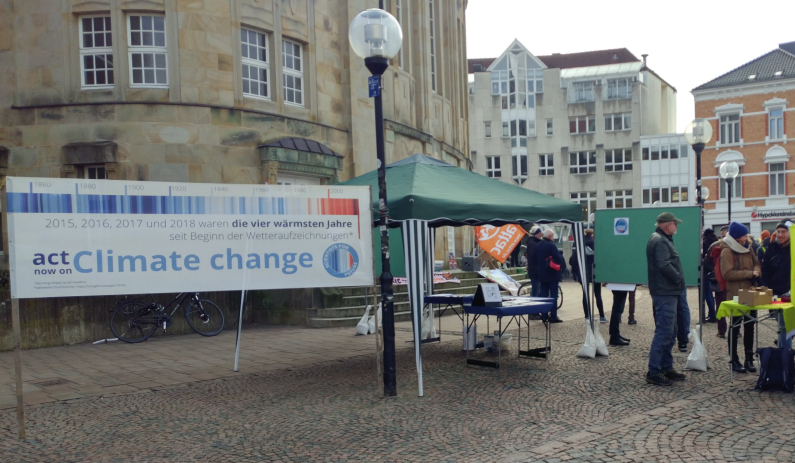 Vor dem Osnabrücker Theater ist ein Banner mit den Klimastreifen gespannt. "act now on Climate change" steht darunter. Daneben das Logo der Scientists for Future. Rechts neben dem Banner steht ein grüner Pavillion. Darunter stehen Tische, auf denen Infomaterial liegt. An einer grünen Stellwand rechts daneben. Mehrere, warm gekleidete Menschen mit Mützen stehen auf dem Platz.