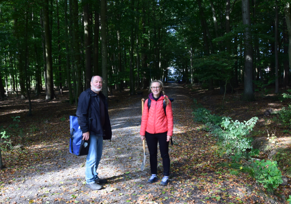 Befestigter gerader Schotterweg im stark belaubten und deshalb dunklen Buchenwald, seitlich Entwässerungsgraben, im Vordergrund eine Frau und ein Mann mit Spaten und Materialtasche, kurz vor Entnahme von Bodenproben am Grieseling