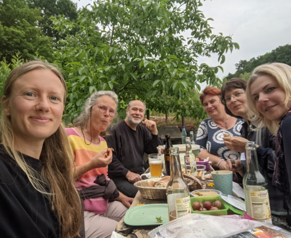 Beim Planungstreffen 375 Friedensbäume sitzen sechs Menschen in einem Garten um einen schmalen Tisch mit allerlei leckerem Essen und Getränken.