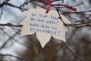 Papier Ahornblatt mit Bastband an einem Zweig befestigt. Darauf steht "Wir sind hier: Wir sind viele! Haltet euch an KLIMAZIELE!"