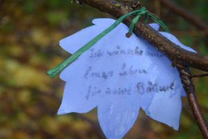 Papier Ahornblatt mit Bastband an einem Zweig befestigt. Darauf steht "Ich wünsche mir langes Leben für unsere Bäume"